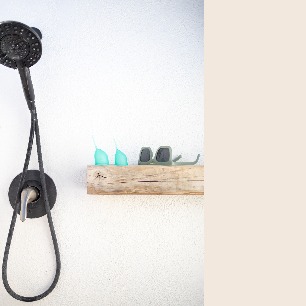Kind Cup menstrual cups in aqua pictured on a wood shelf next to a black shower fixture and sunglasses .