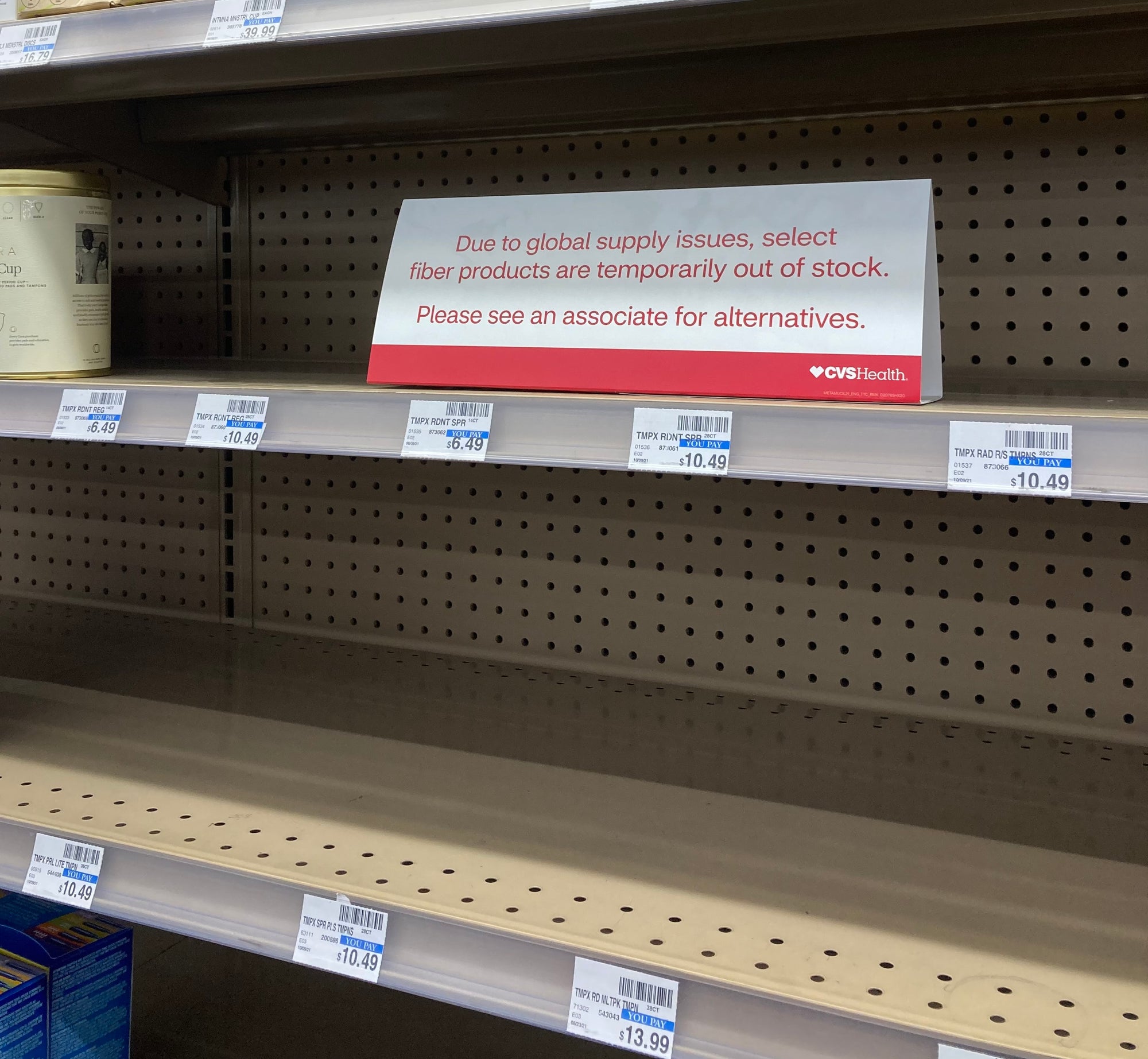 Image of sign on CVS period product shelf stating there's a global shortage impacting availability of tampons. Metallic shelves are largely empty.