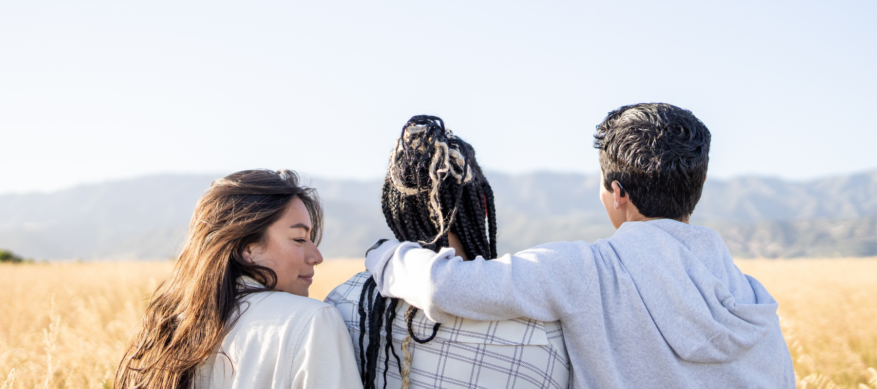 Three people of different races stand closely together in a comfortable embrace in a beautiful open field of dried grasses with mountains in the distance.