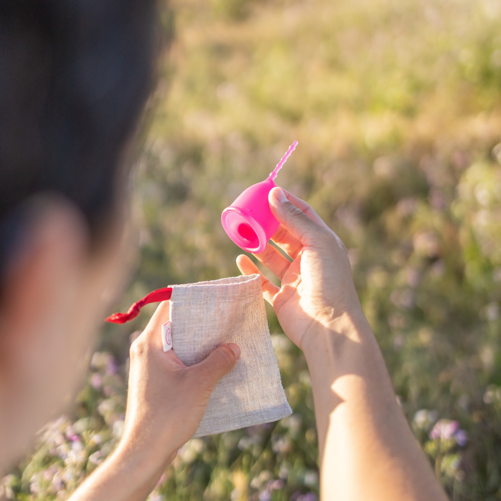 Person looking at Kind Cup size regular in violet menstrual cup and holding it above the earth-friendly linen bag in a field of wildflowers.